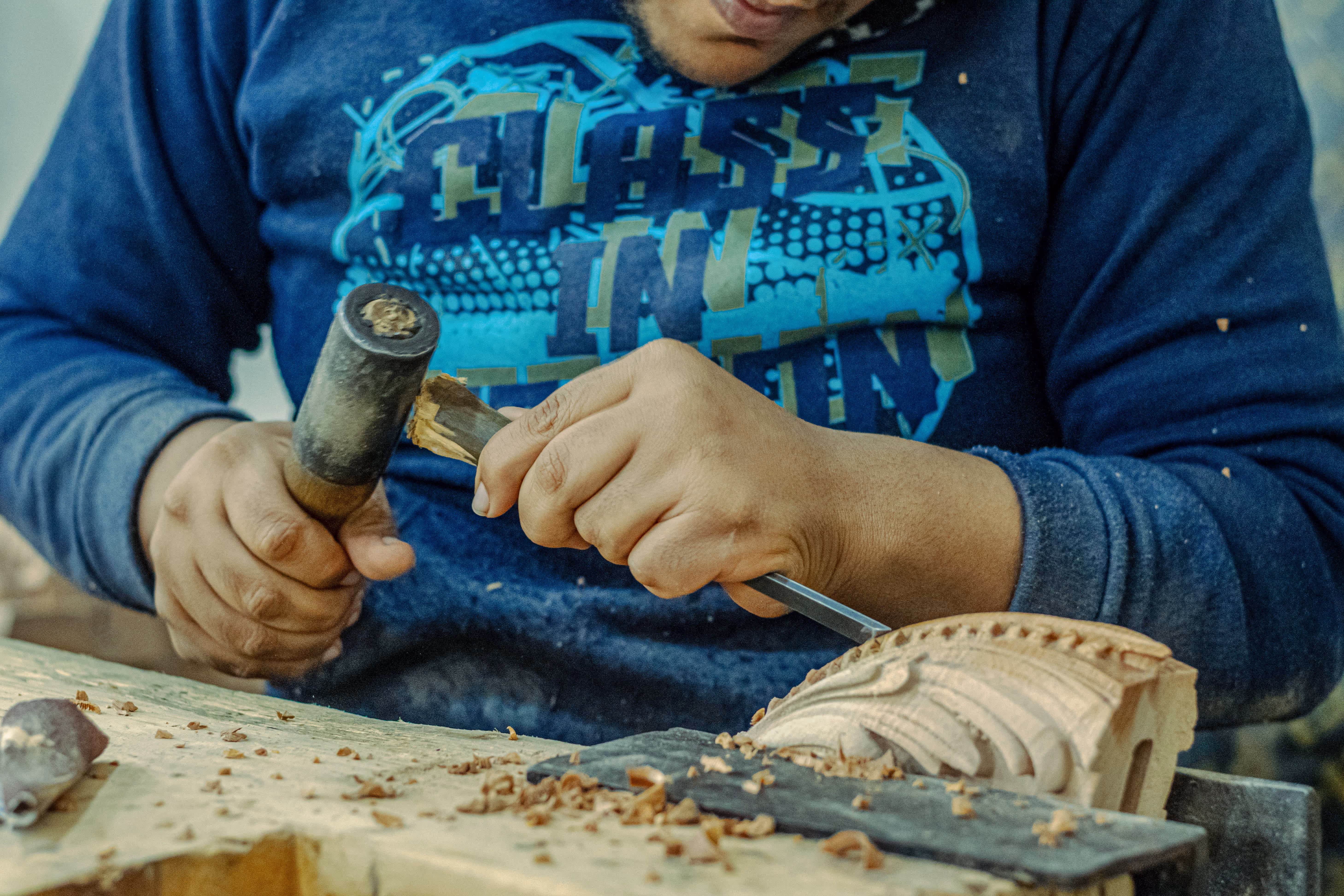 Person in blue sweatshirt carving detailed wood design using chisel and mallet.