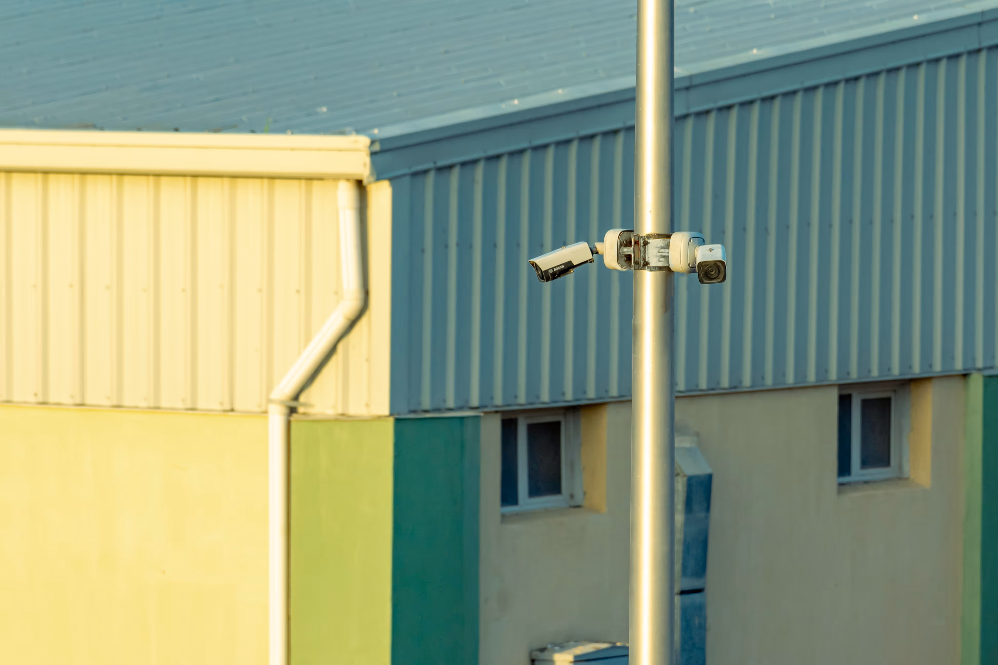 Two security cameras mounted on a pole in front of a building with blue and yellow walls.