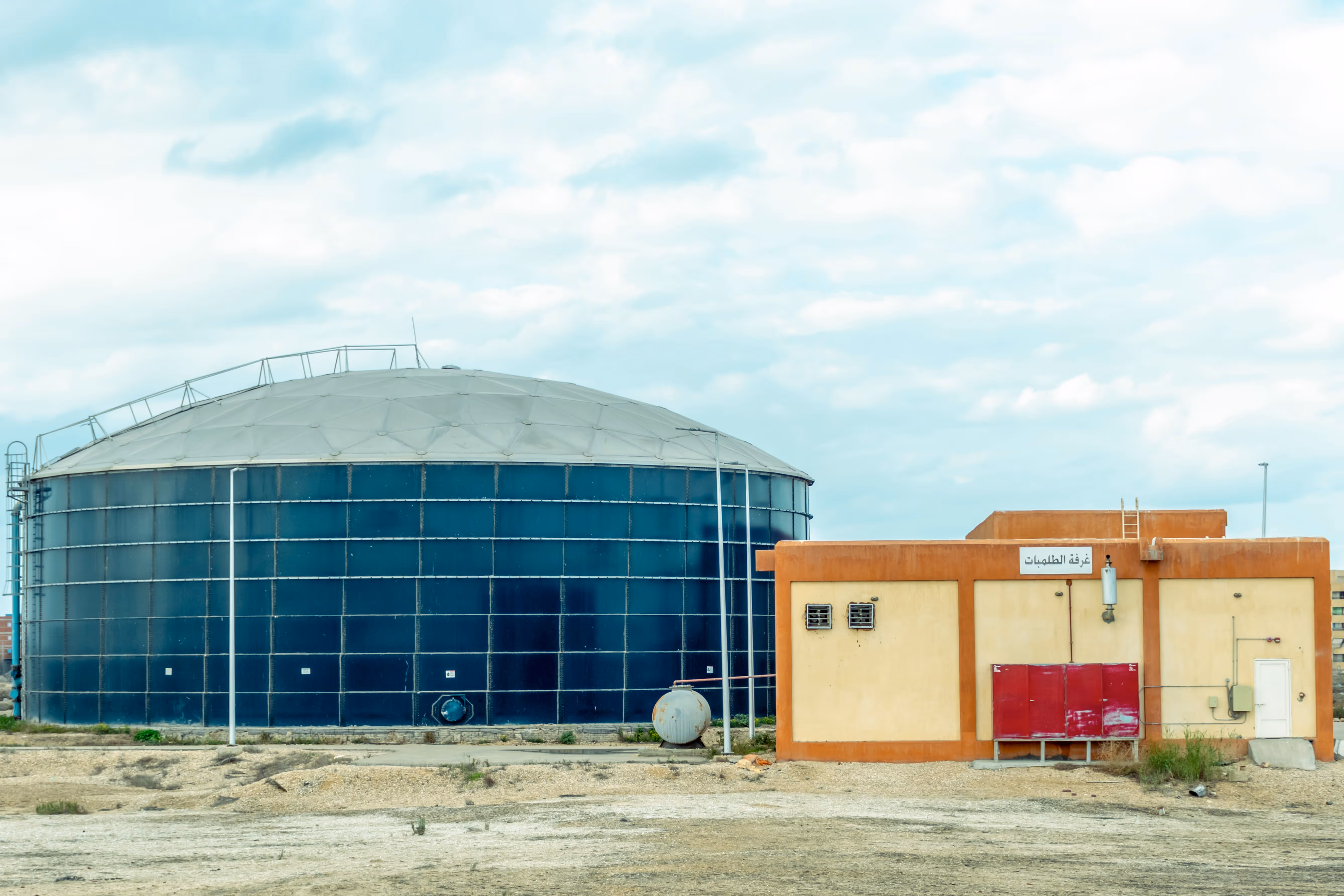 Large blue industrial storage tank with dome roof next to a beige building with red panels.