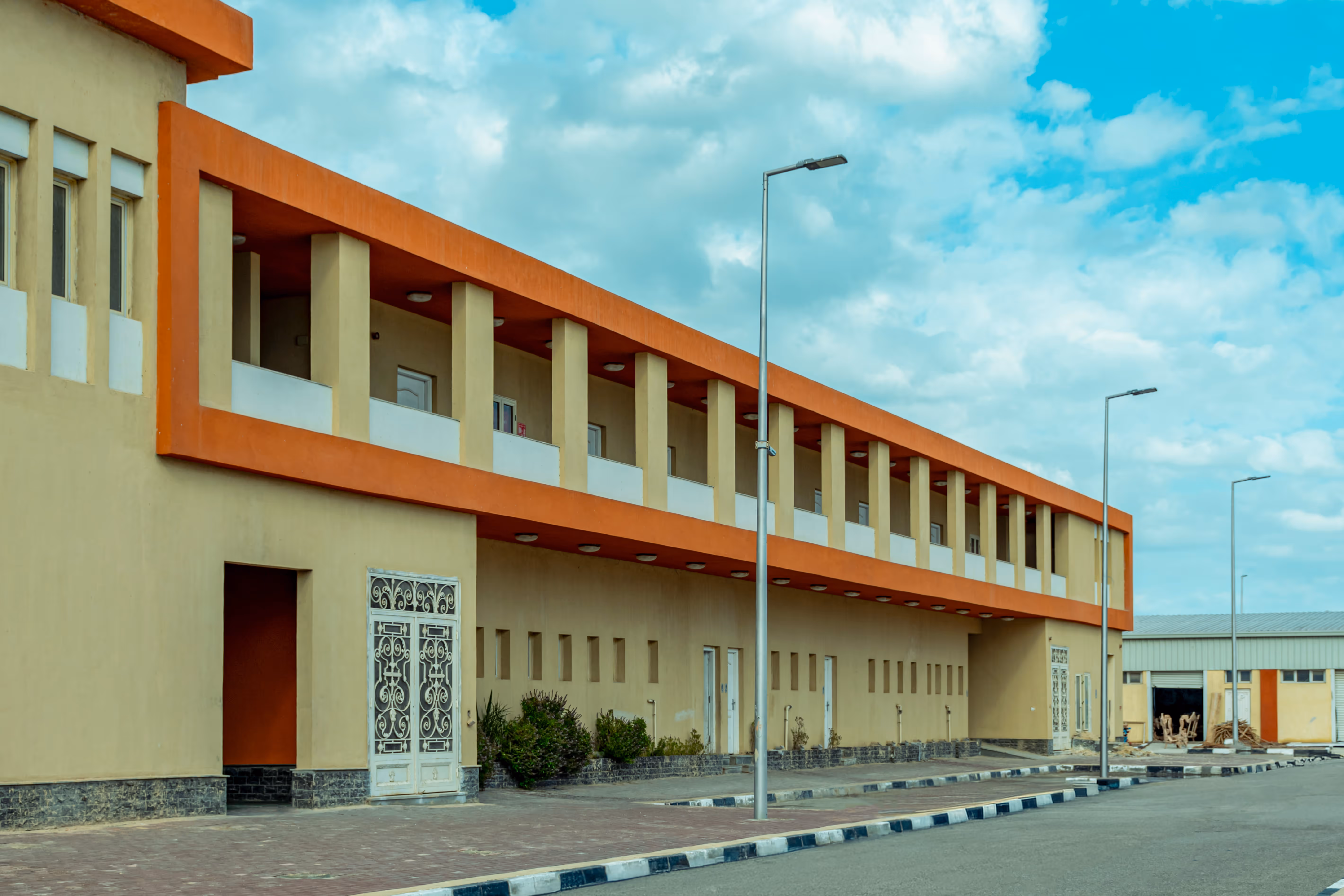 Long beige building with orange trim and pillars under a partly cloudy blue sky.