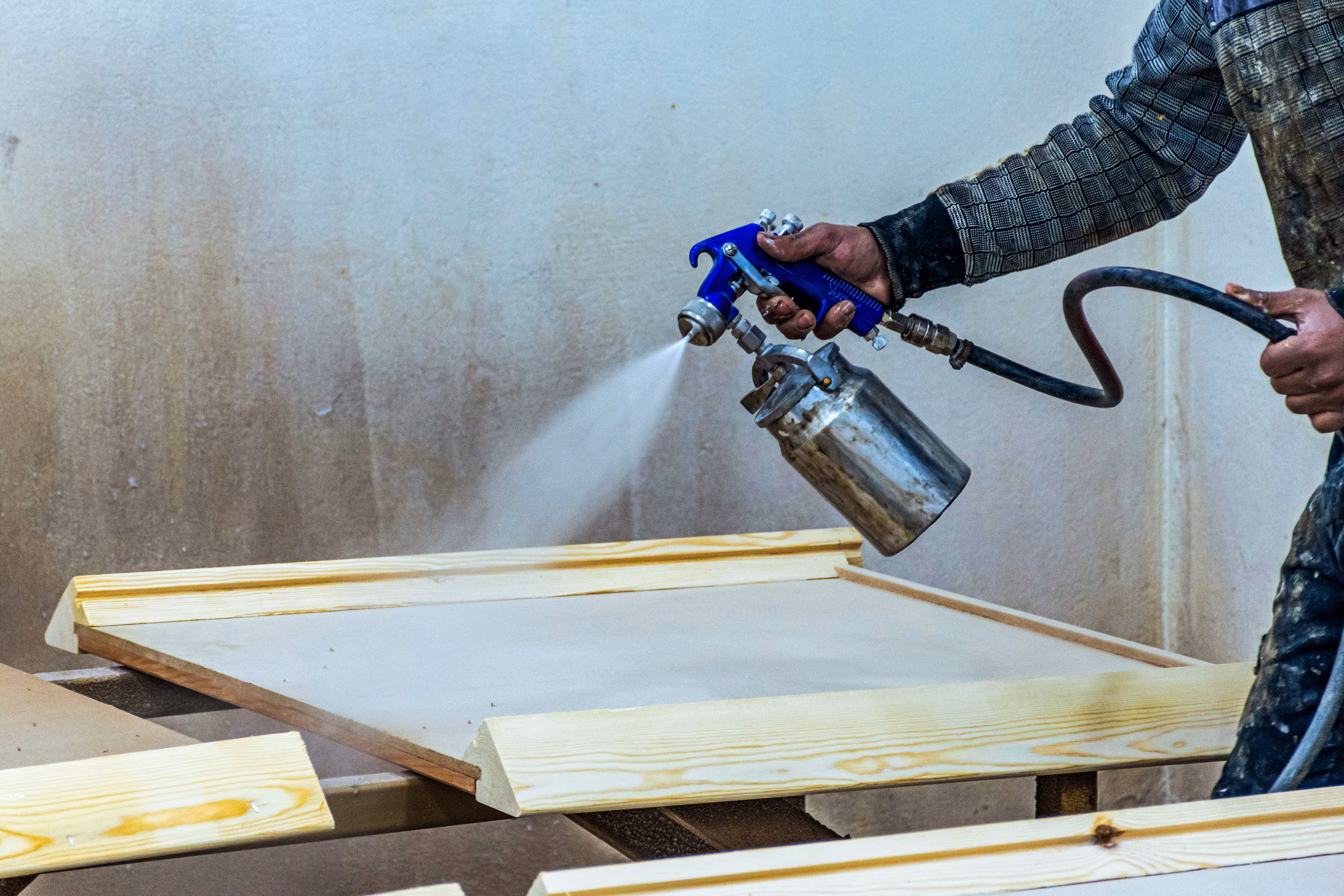 Person spraying wood panel with a spray gun in a workshop.