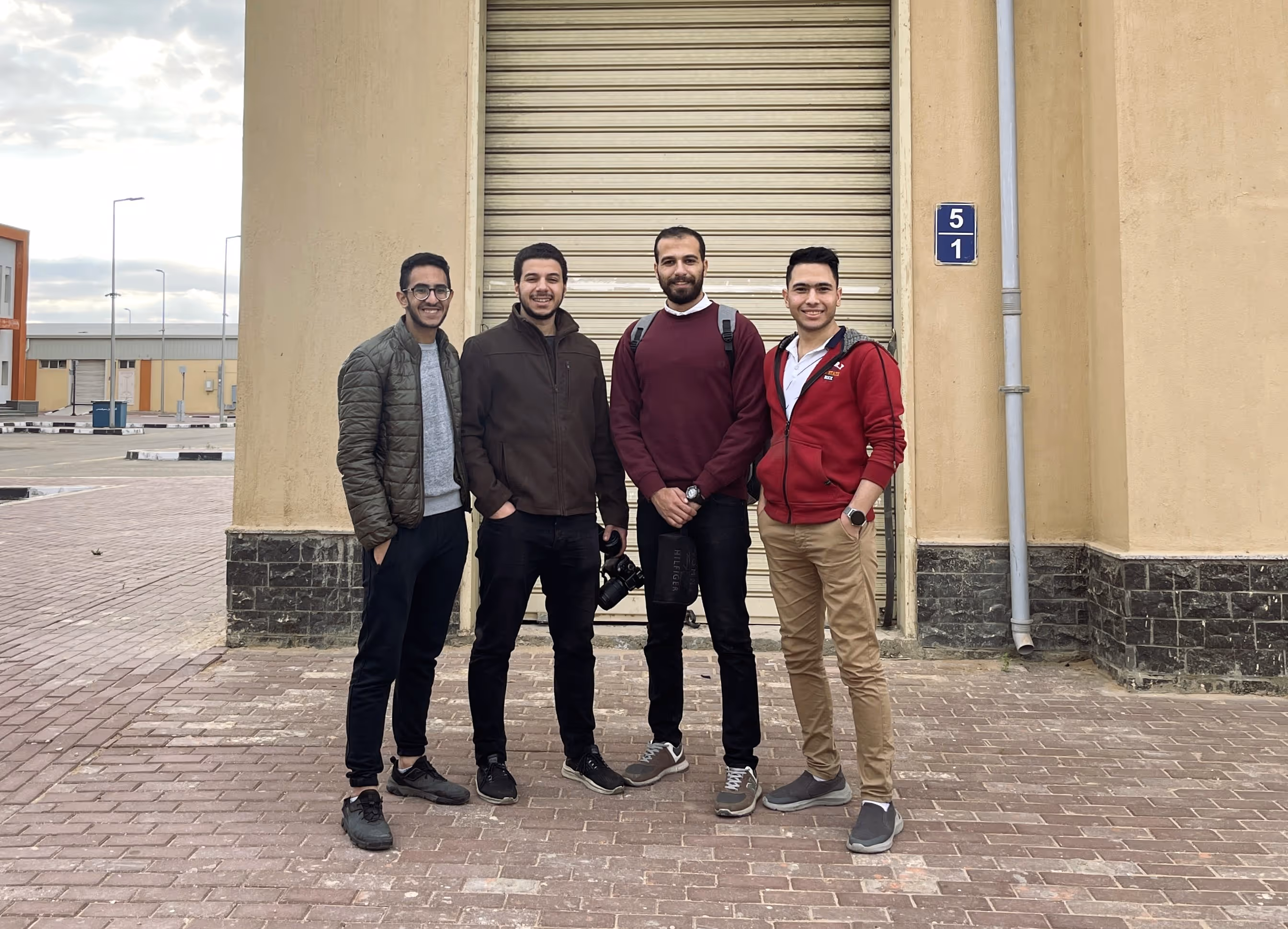 Four young men standing on a brick path in front of a beige building with a closed shutter.