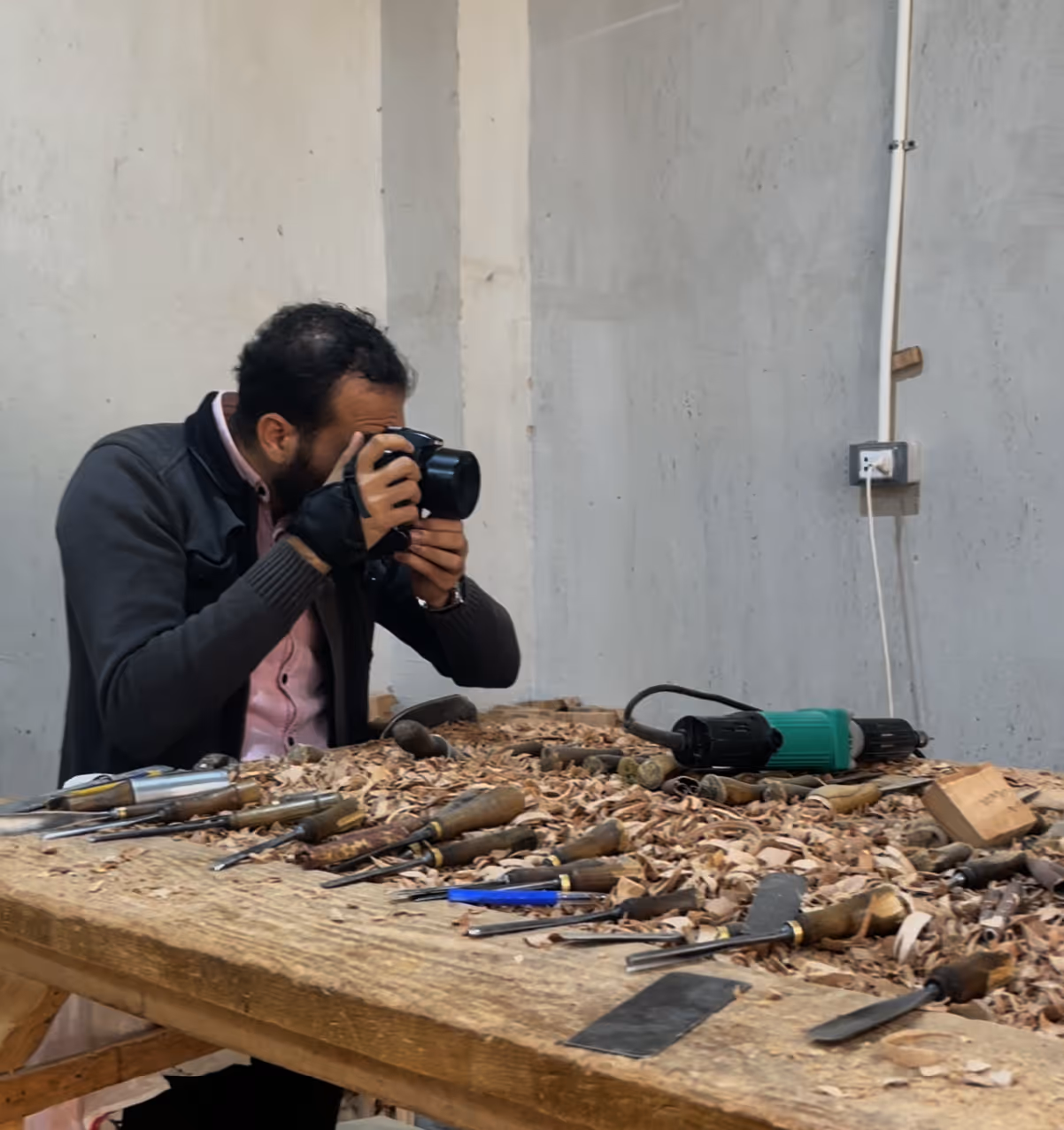Man with camera photographing a woodshop table covered in wood shavings and carving tools.