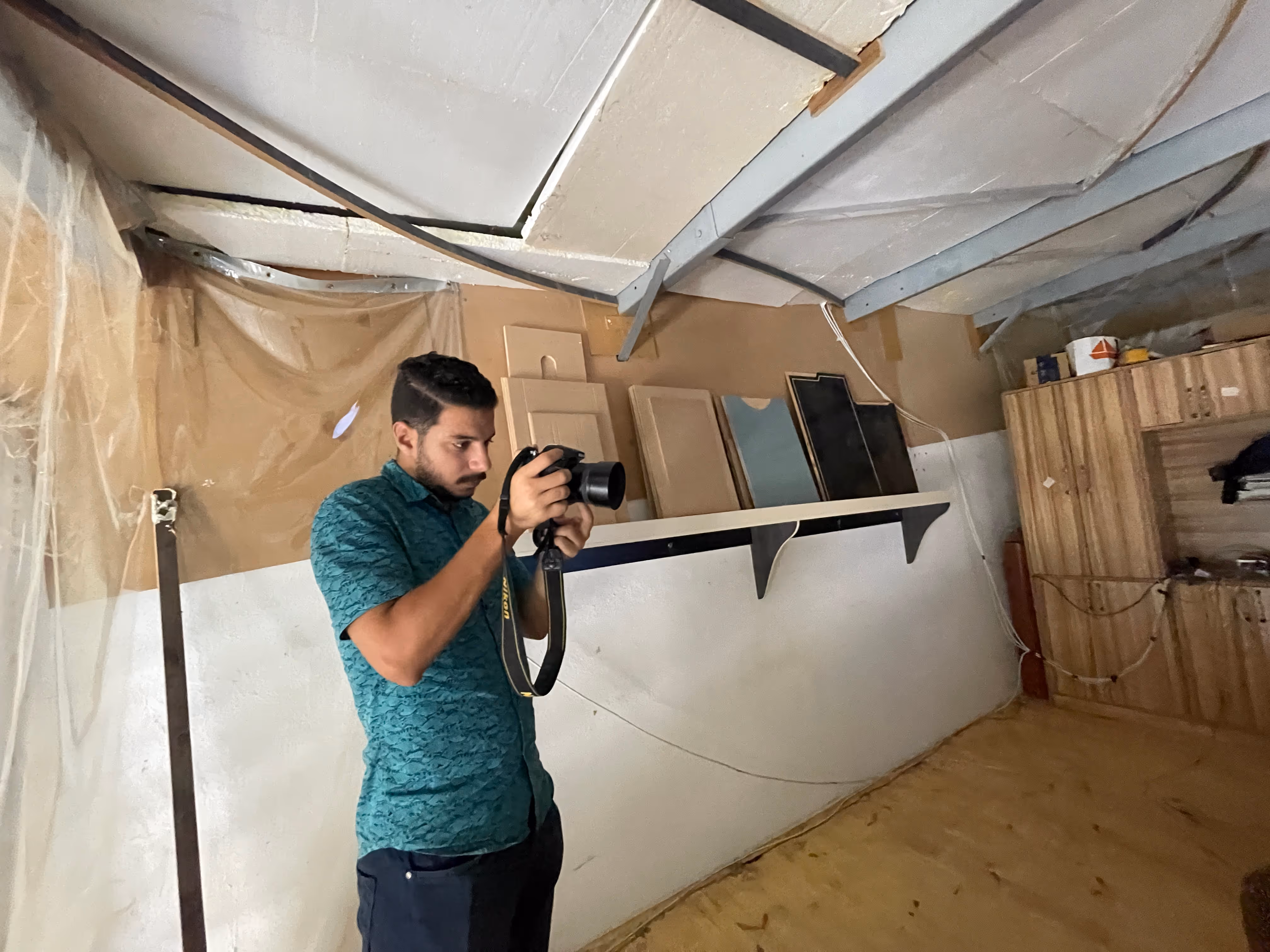 Man in teal shirt holding a camera in a room with unfinished ceiling and wood panels on the wall.