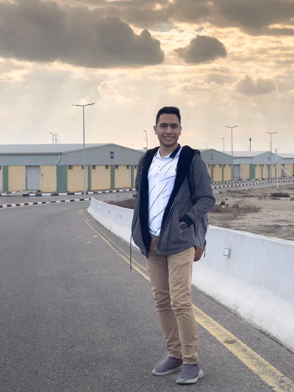 Man in a gray jacket and beige pants standing on a road with industrial buildings and cloudy sky.