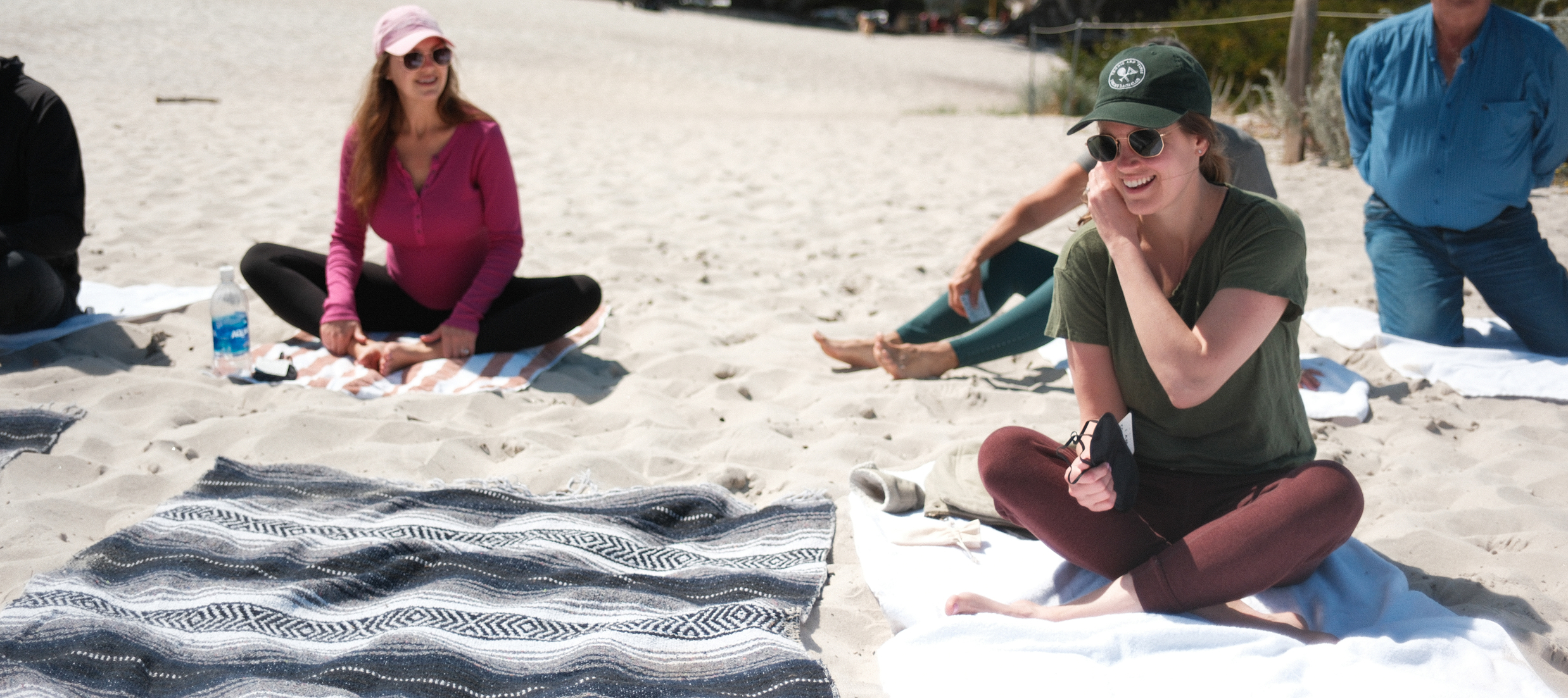 Wedding guests sitting on towels in the sand, smiling and talking before a beach breathwork session