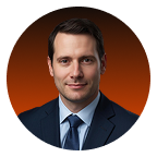 Portrait of a man in a dark suit and tie with a neutral expression against a dark orange background.