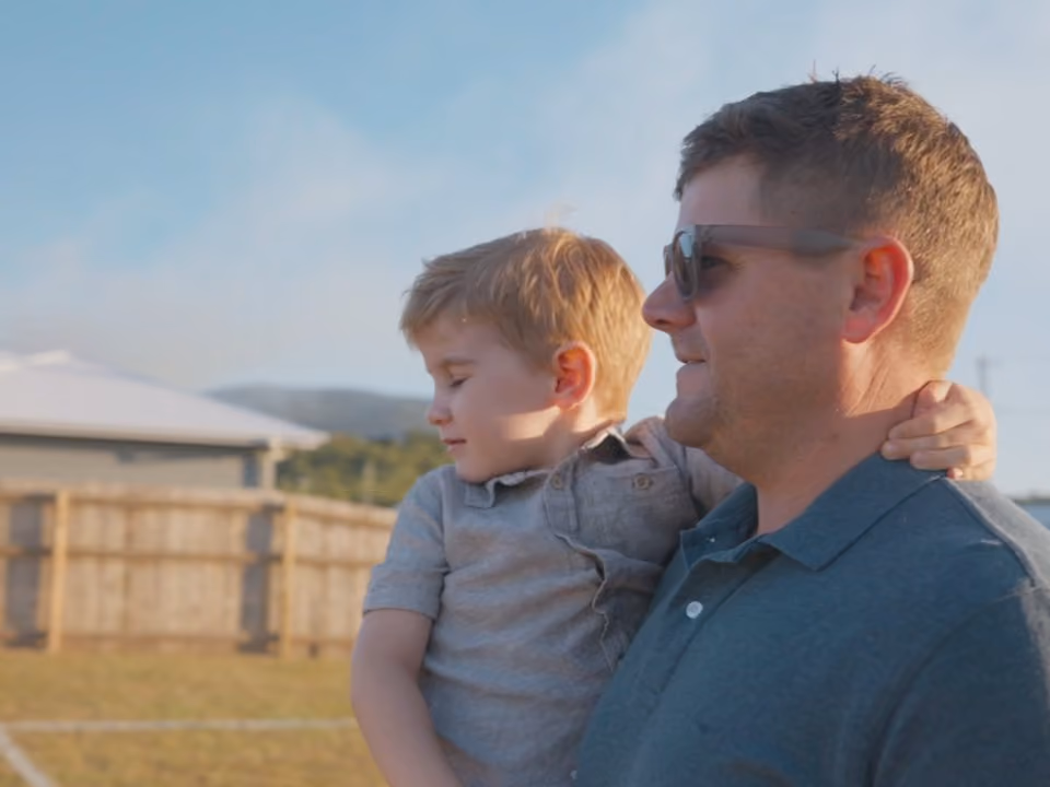 Man wearing sunglasses holds a young boy outside near a wooden fence under a blue sky.
