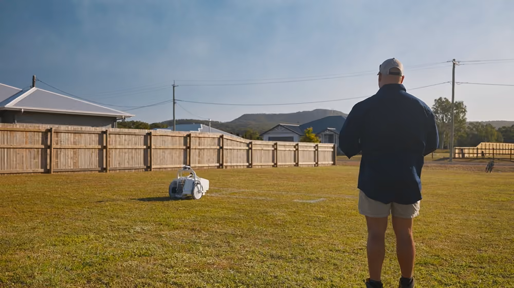 Man standing on a grassy field controlling a white remote-controlled vehicle near a wooden fence.