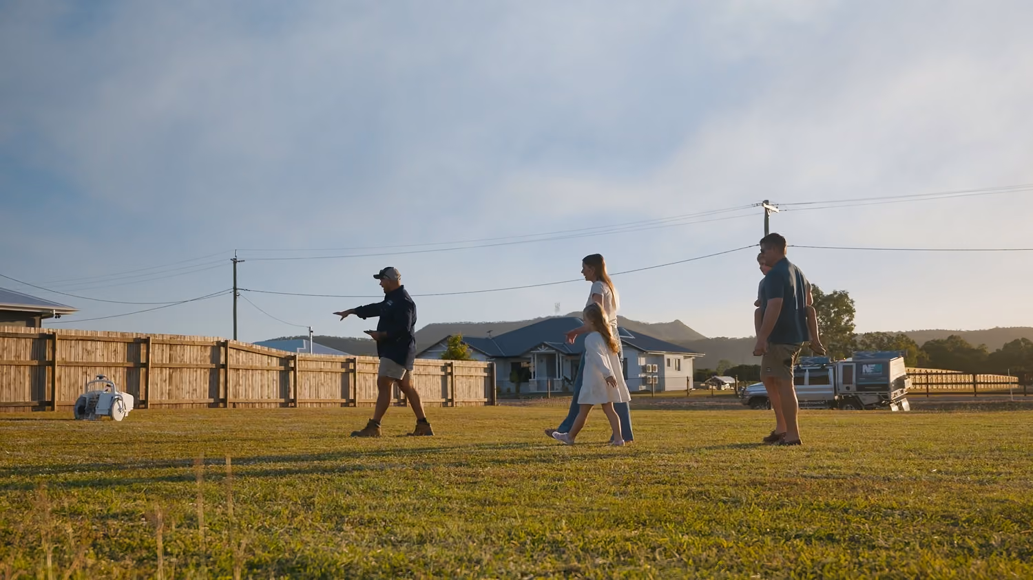 Five people walk on a grassy lawn near a wooden fence and house with mountains in the background.