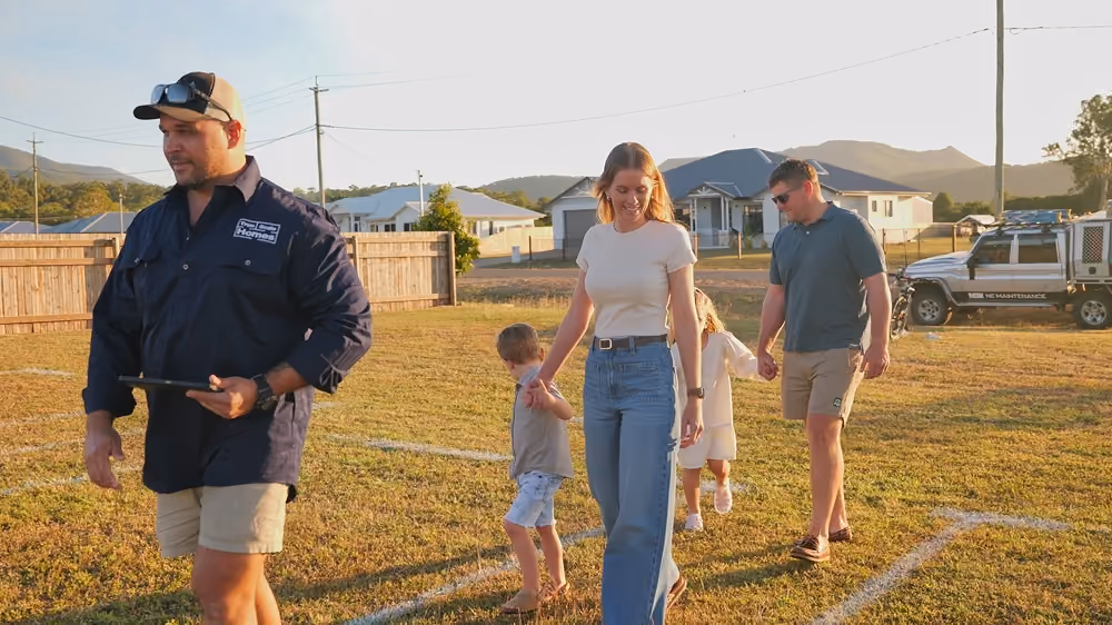 Man in branded shirt walks ahead of a family of four holding hands on a grassy lot with house outlines.