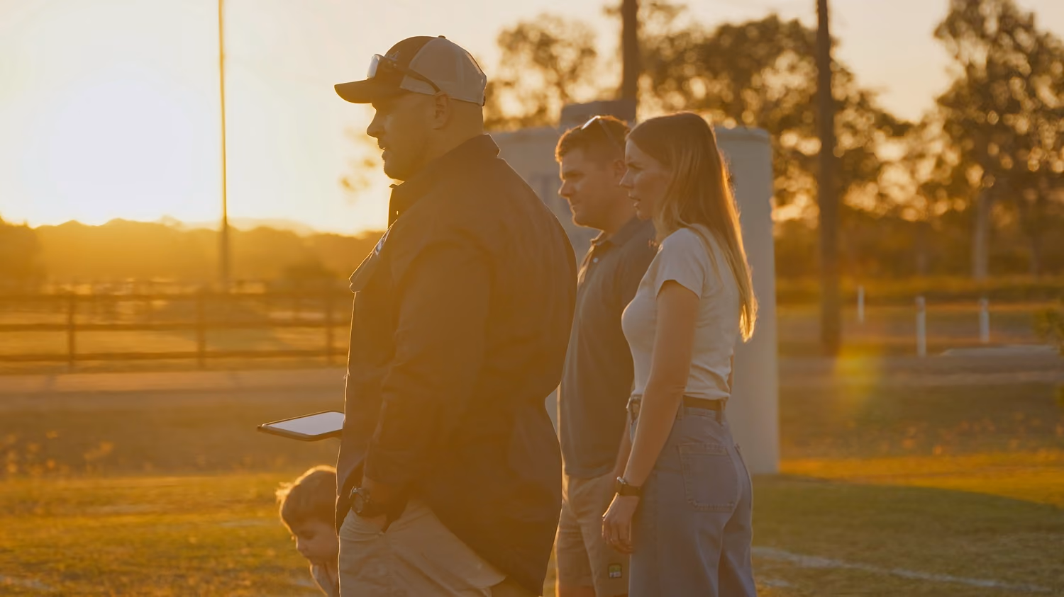 Three adults and a child standing outdoors at sunset in a rural area with trees and fences.