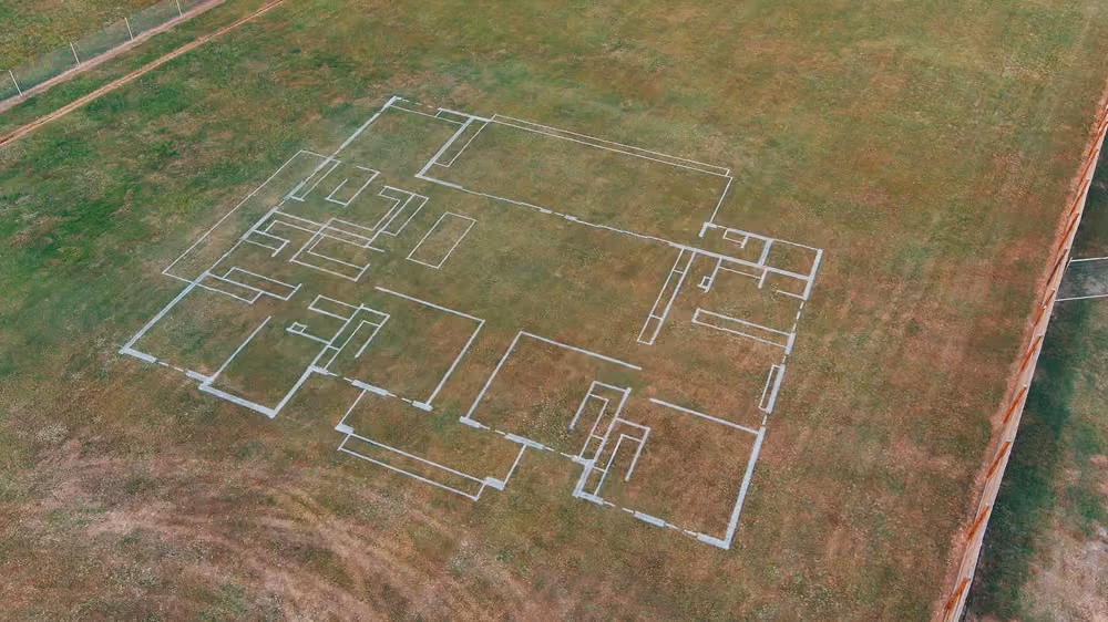 Outline of a house floor plan marked with white lines on a grassy ground, viewed from above.