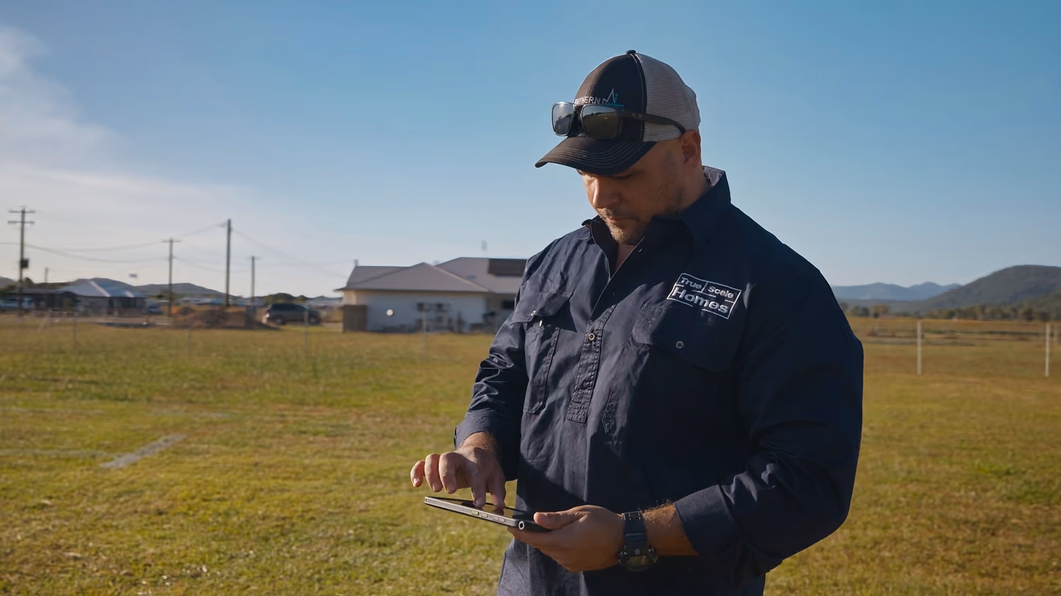 Man in a cap and True Scale Homes shirt using a tablet outside on a sunny day with houses in the back.