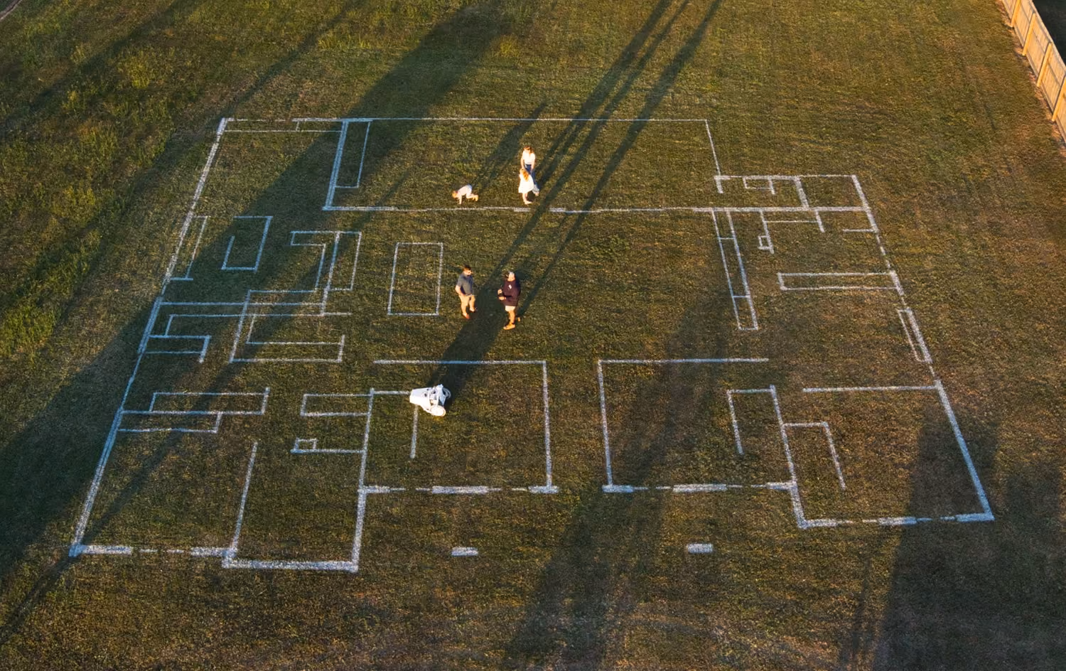 Aerial view of a house floor plan marked on grass with four people and a dog standing inside the layout.