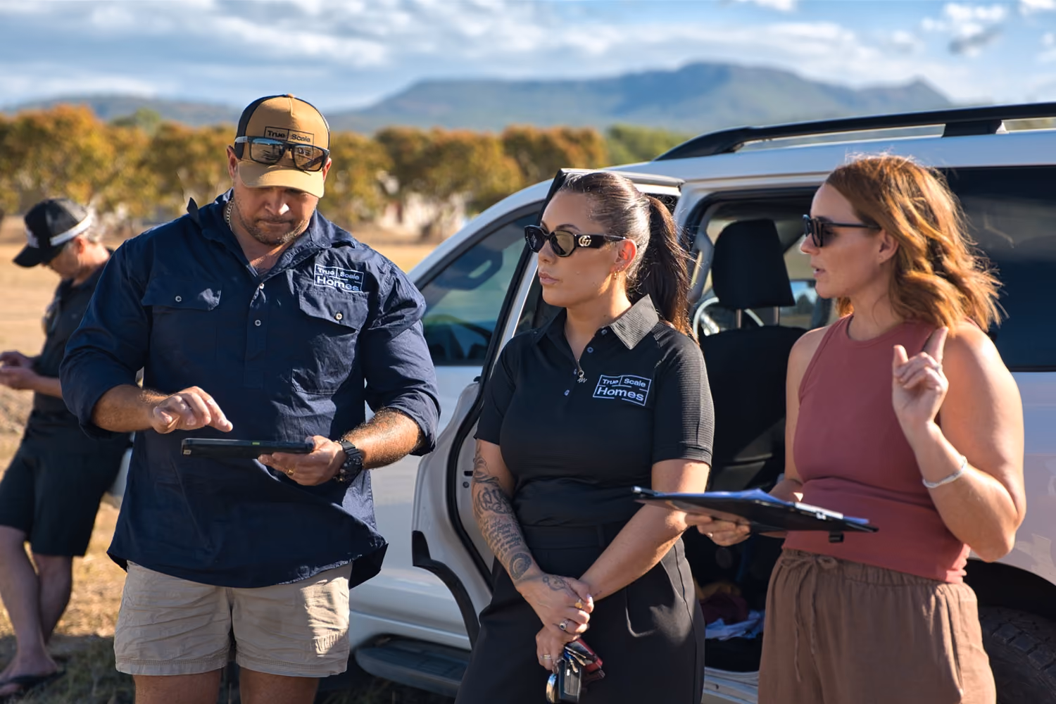 Three people talking near a white SUV; two wear True Scale Homes shirts, one holds a clipboard.