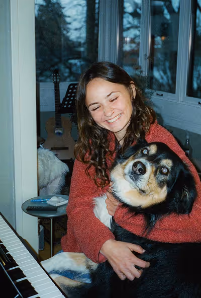 Smiling woman in a red sweater hugging a large black and tan dog indoors near a piano.