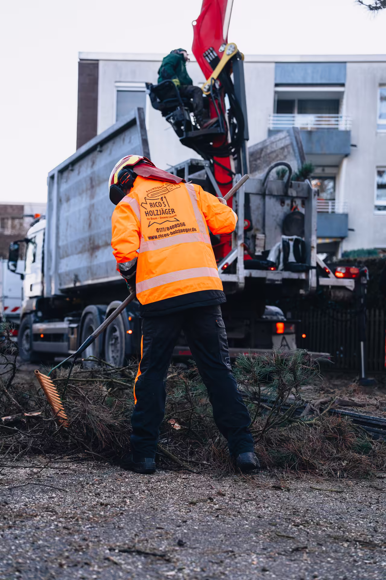 Arbeiter in oranger Sicherheitsjacke sammelt Holzabfälle vor einem Lastwagen mit Kran.