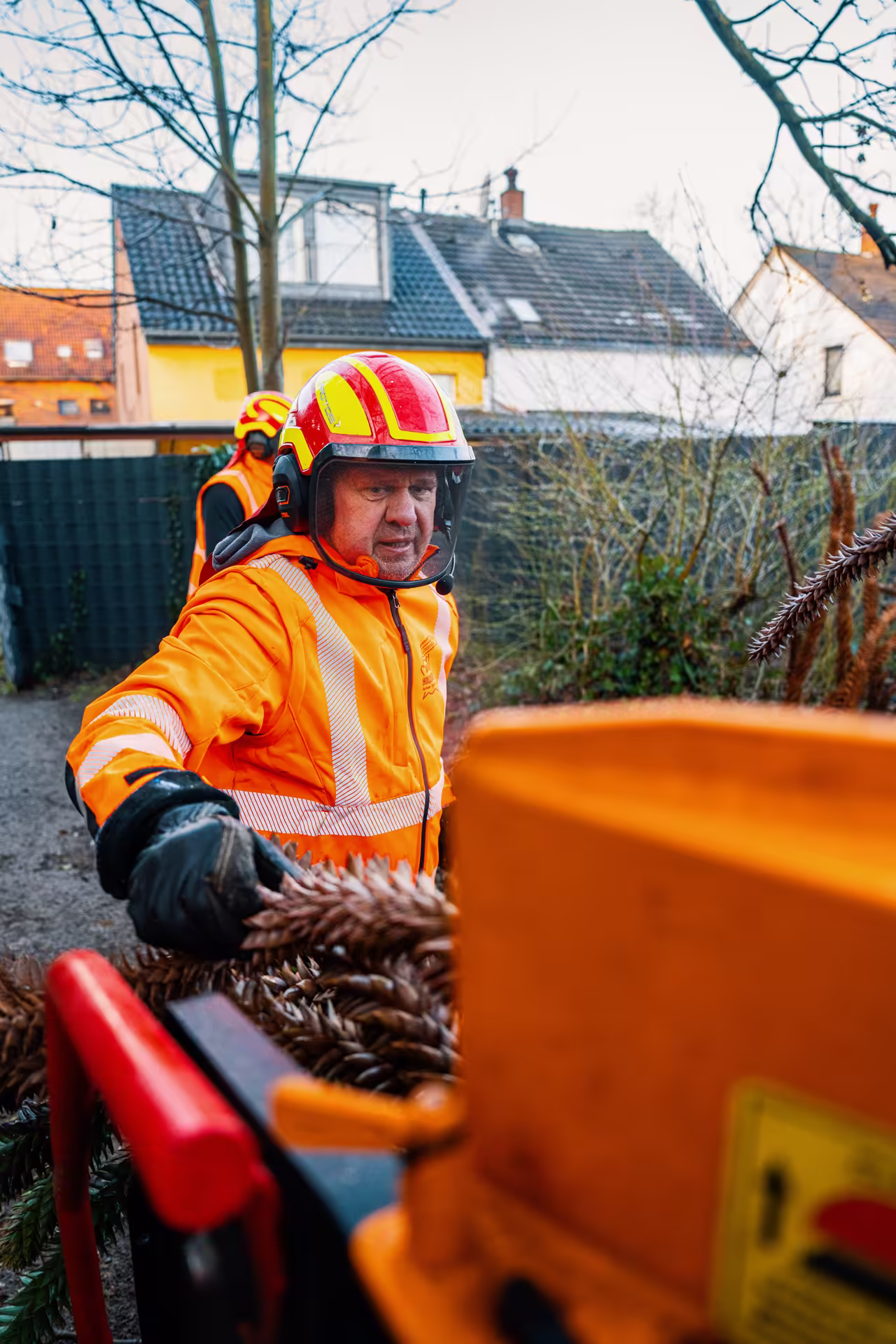 Arbeiter in orangefarbener Schutzkleidung und Helm bedienen Holzschredder im Freien.