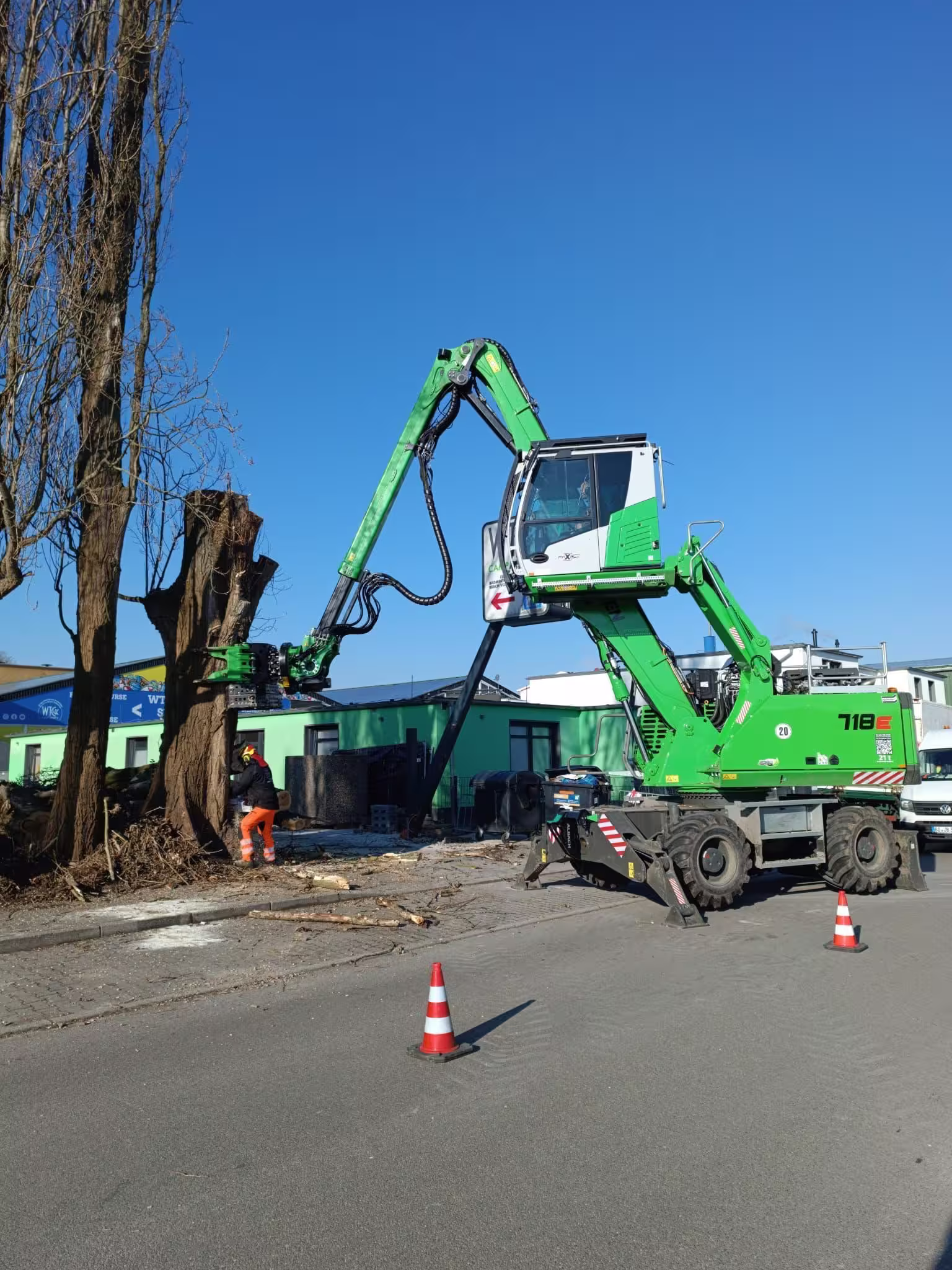 Grüne Baumaschine schneidet Baumstamm auf Straße, Arbeiter mit Schutzkleidung und Verkehrshütchen.