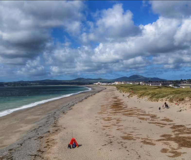 Wide sandy beach with scattered seaweed, a person sitting under a red umbrella, people walking near dunes, ocean on the left, and distant hills under a partly cloudy sky.