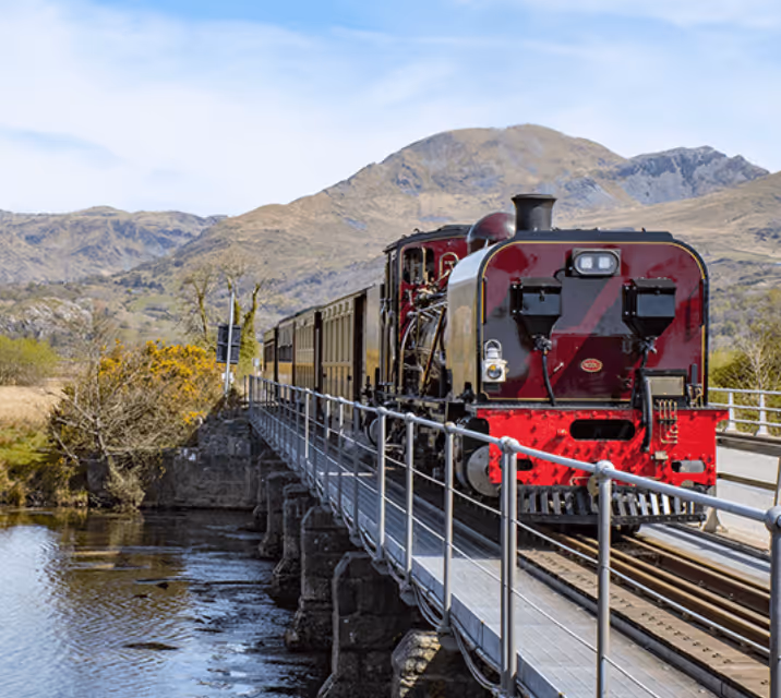 Red and black steam locomotive crossing a bridge over water with mountains in the background.
