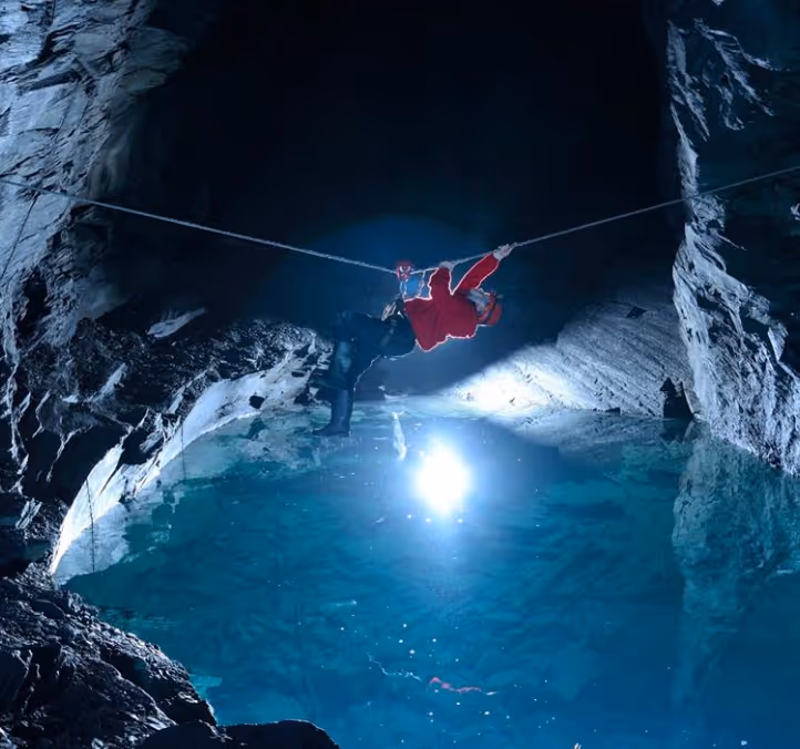 Person in red jacket suspended on a zipline over a clear blue underground cave pool.