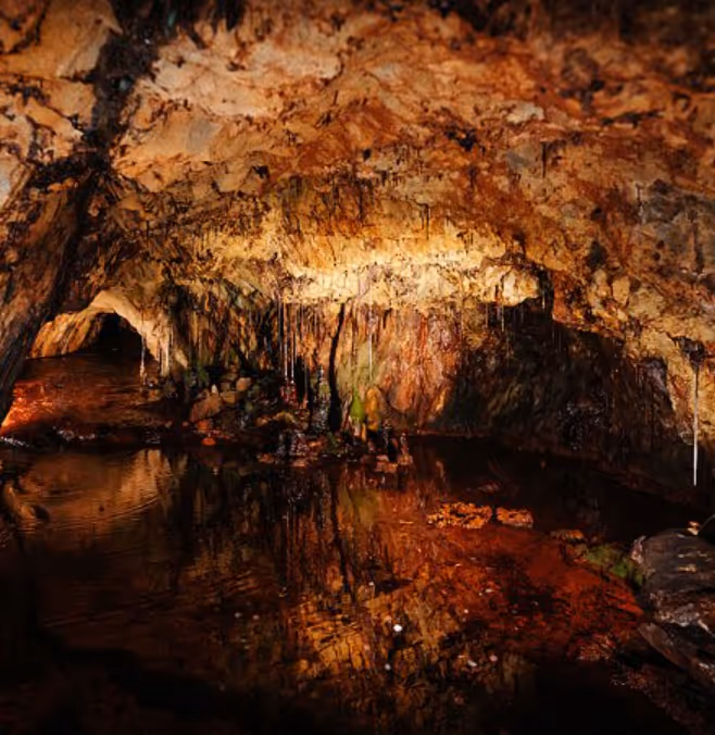 Interior of a dimly lit cave with stalactites hanging from the ceiling and their reflection in the water below.