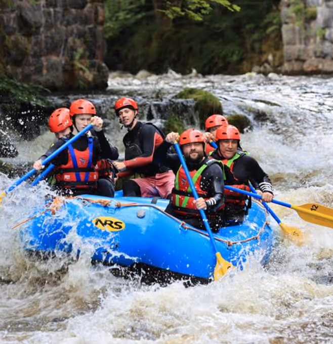 Group of six people wearing helmets and life jackets navigating a blue raft through white water rapids in a river.
