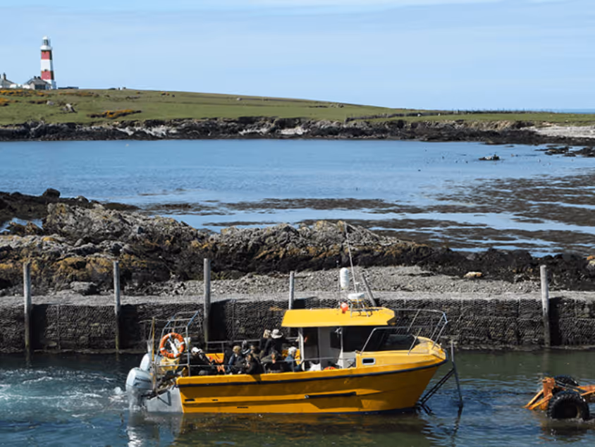 Yellow boat with people on board near a rocky shoreline and a red-and-white lighthouse in the background.