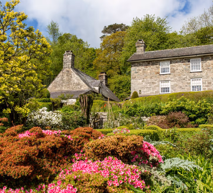 Stone cottages surrounded by lush greenery and colorful blooming flowers under a partly cloudy sky.