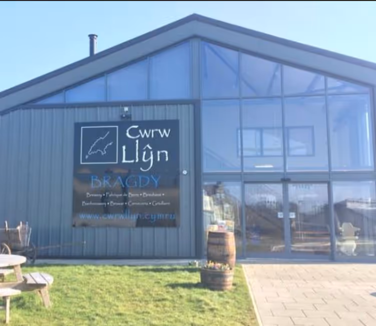 Gray building with large glass windows displays a sign for Cwrw Llŷn brewery and pub on a sunny day with green grass and wooden barrels in front.