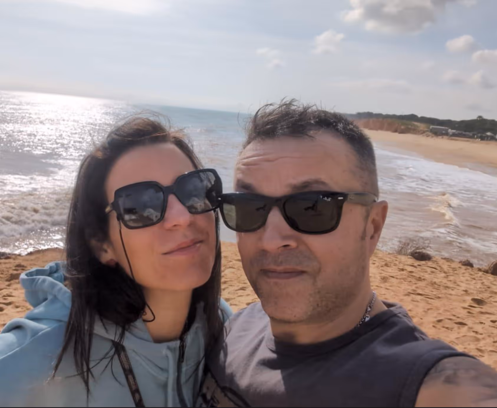 Man and woman wearing sunglasses taking a selfie at a sunny beach with waves and a partly cloudy sky.
