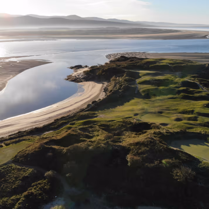 Aerial view of a golf course by a river estuary with sand dunes and distant mountains under a bright sky.