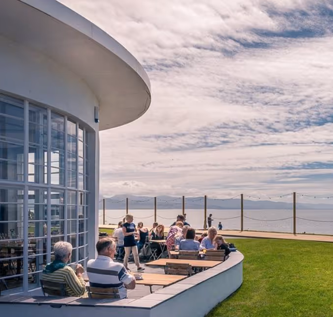 People sitting and standing at outdoor tables beside a white curved building overlooking a body of water with mountains and a partly cloudy sky in the background.