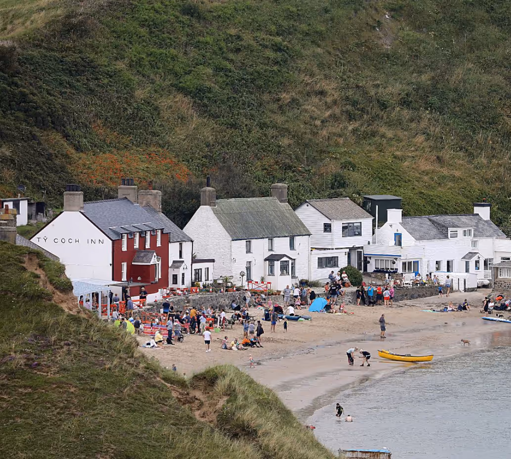 Crowded sandy beach in front of white and red houses including the Ty Coch Inn, with green hillside in the background.