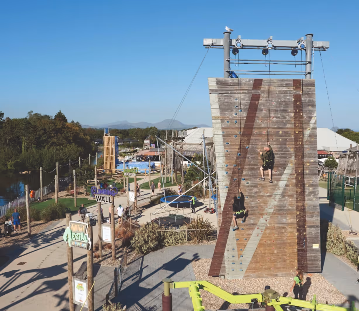 Two people climbing a wooden rock climbing wall outdoors with a clear blue sky and activity park in the background.