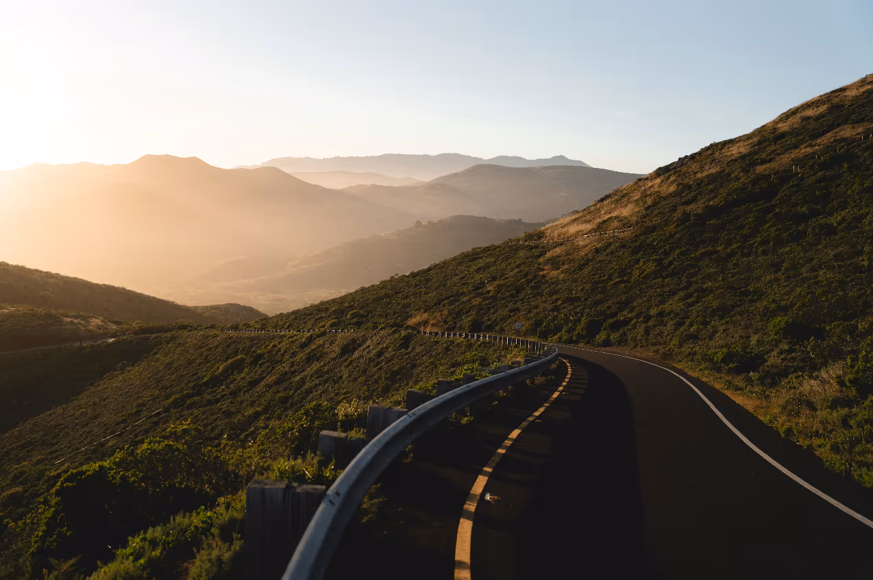 Winding road through green hills with soft sunlight and distant mountain layers under clear sky.