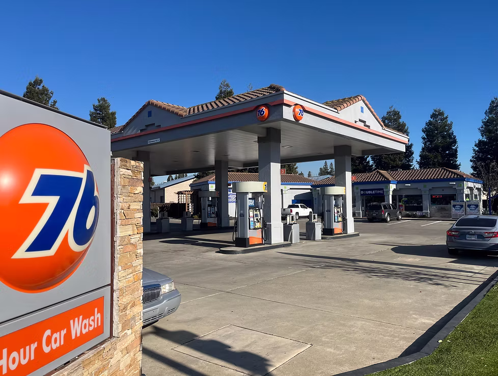 76 gas station with multiple fuel pumps under a canopy and a sign advertising a 24-hour car wash on a clear sunny day.