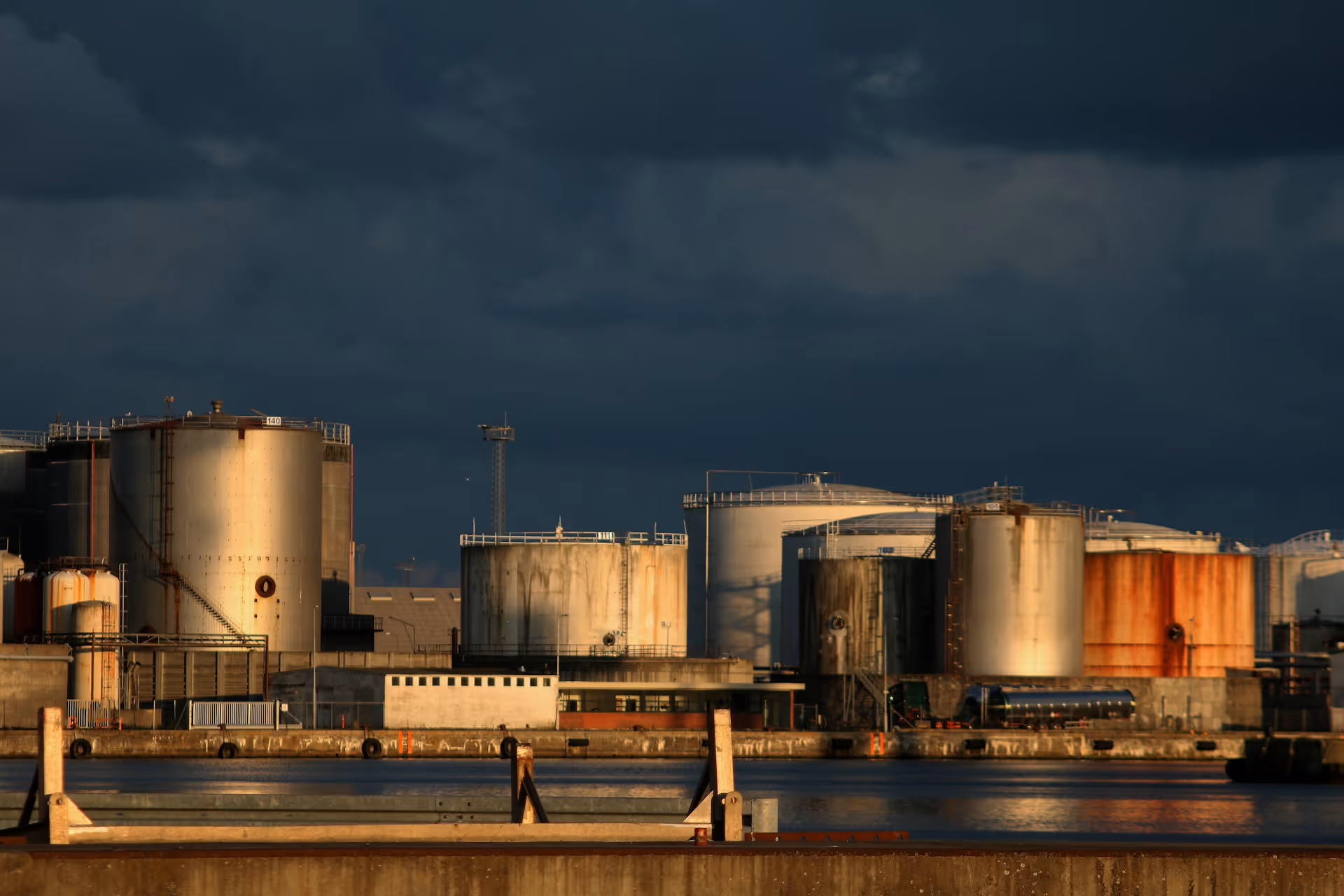 Industrial storage tanks at a port under dramatic dark clouds with sunlight reflecting off the tanks.