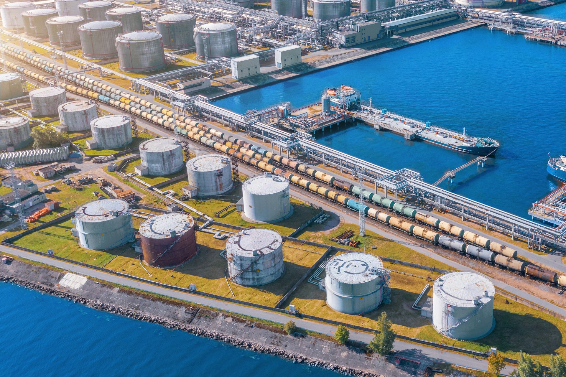 Aerial view of an industrial port with large cylindrical storage tanks, tanker ships docked, and a rail line with tank cars.