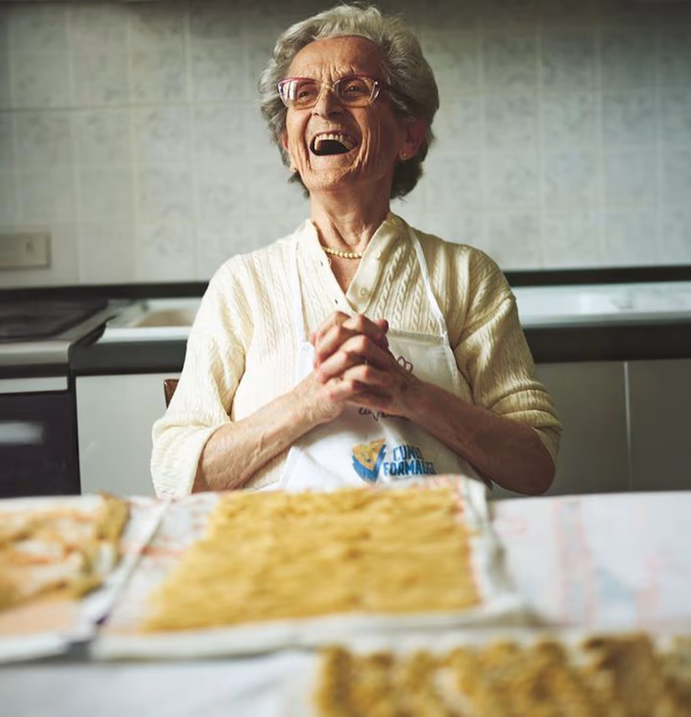 Elderly woman wearing glasses and apron, smiling and clasping hands in a kitchen with pasta on the table.