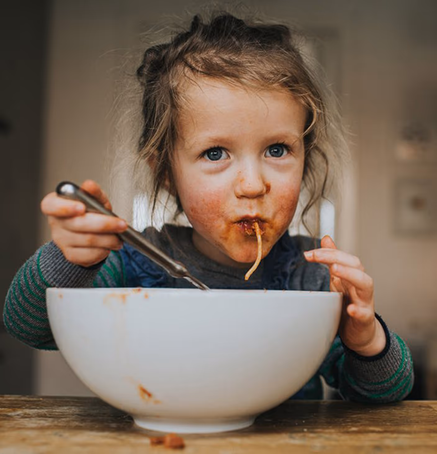 Young girl with messy hair and a sauce-stained face eating spaghetti from a large white bowl.