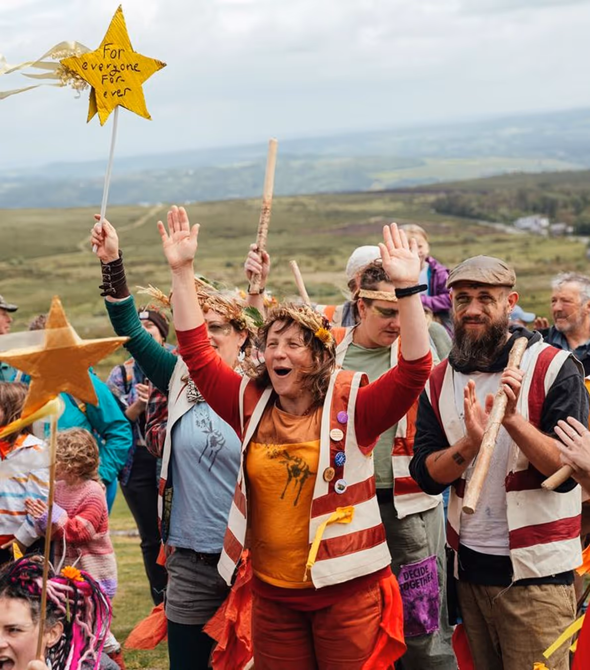 Group of people outdoors celebrating, some raising hands and holding star-shaped wands, one wand reads 'For everyone for ever'.