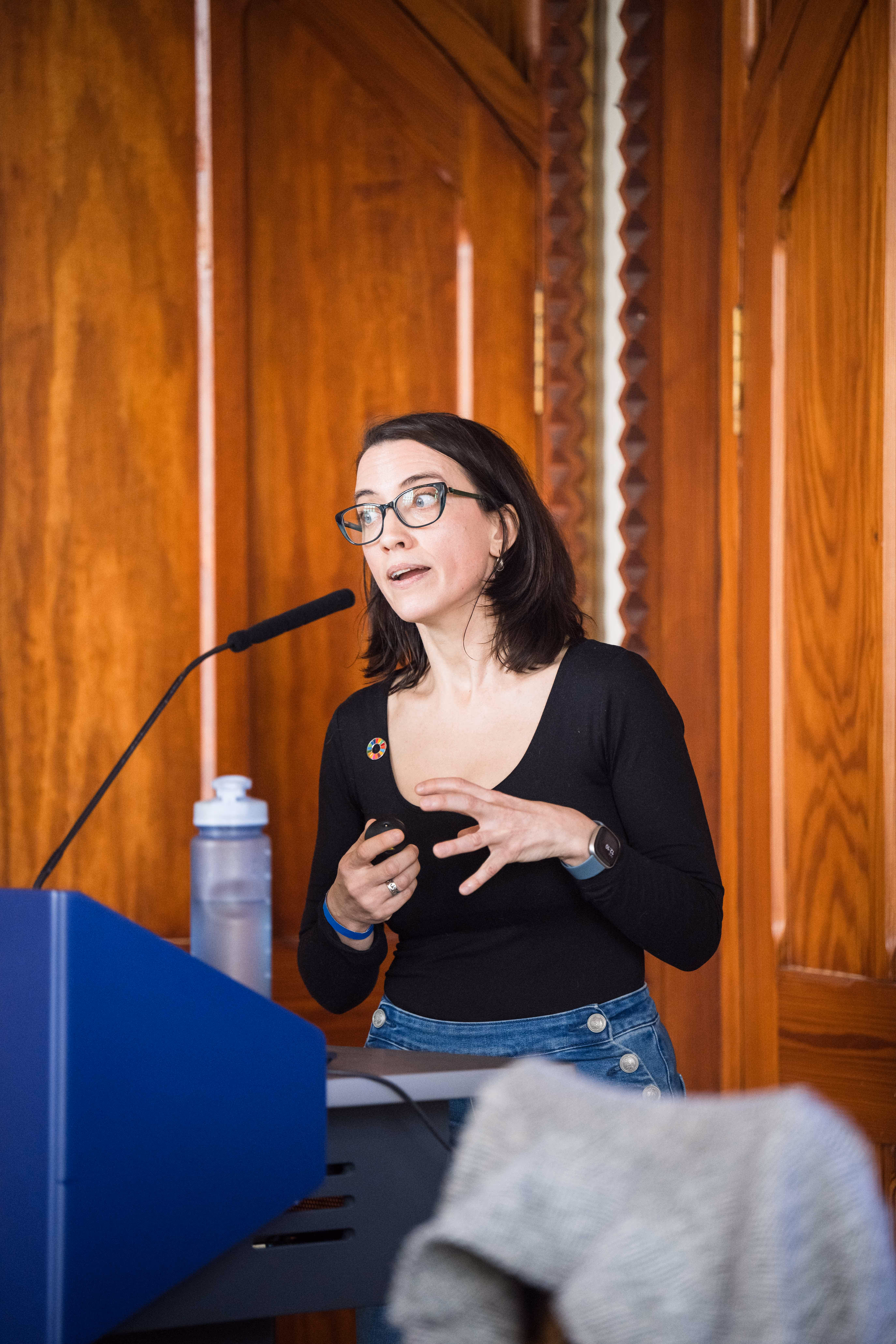 Woman with glasses and dark hair speaking at a podium with a microphone in a wood-paneled room.