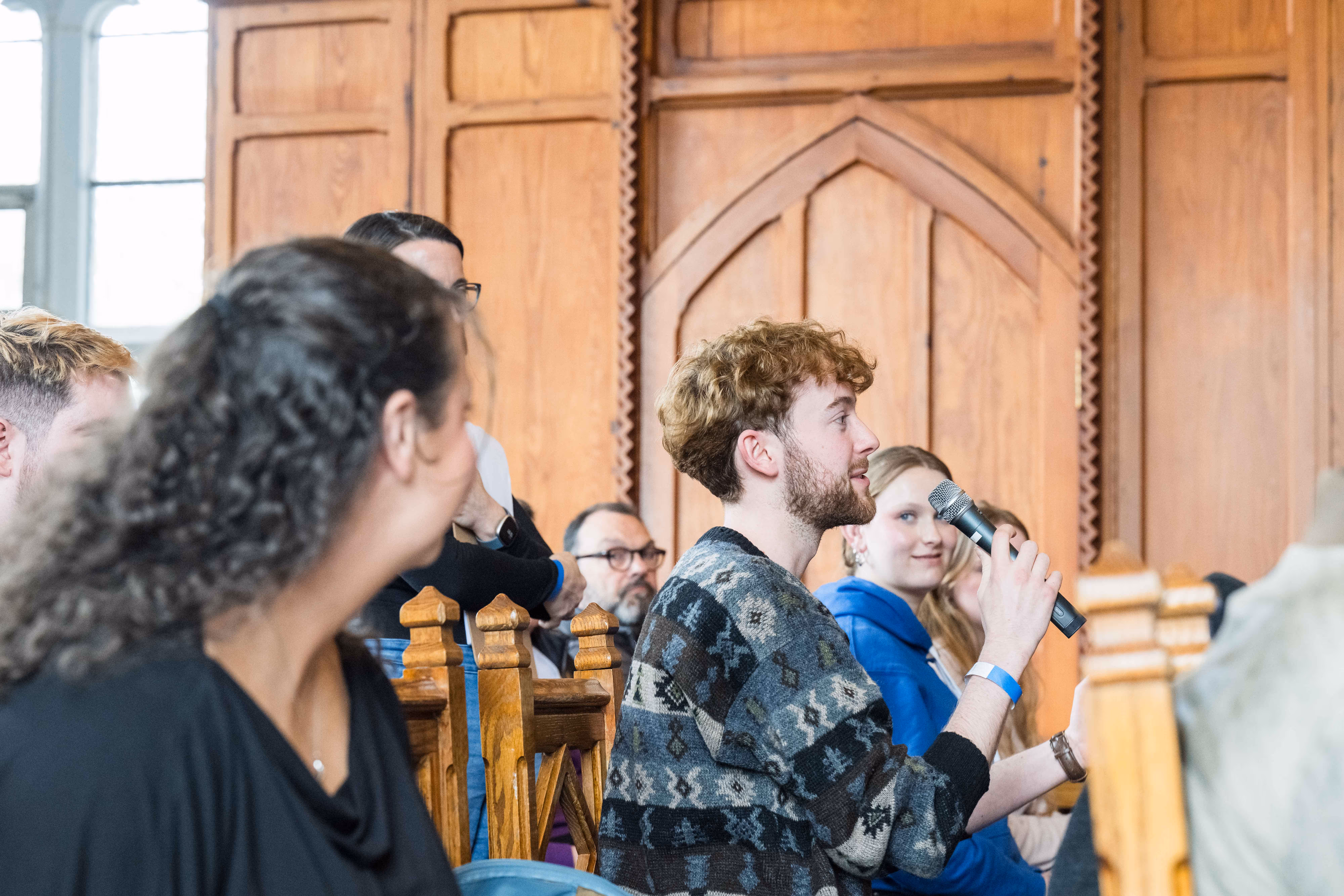 Man with curly hair speaking into a microphone during a seated group discussion in a wood-paneled room.