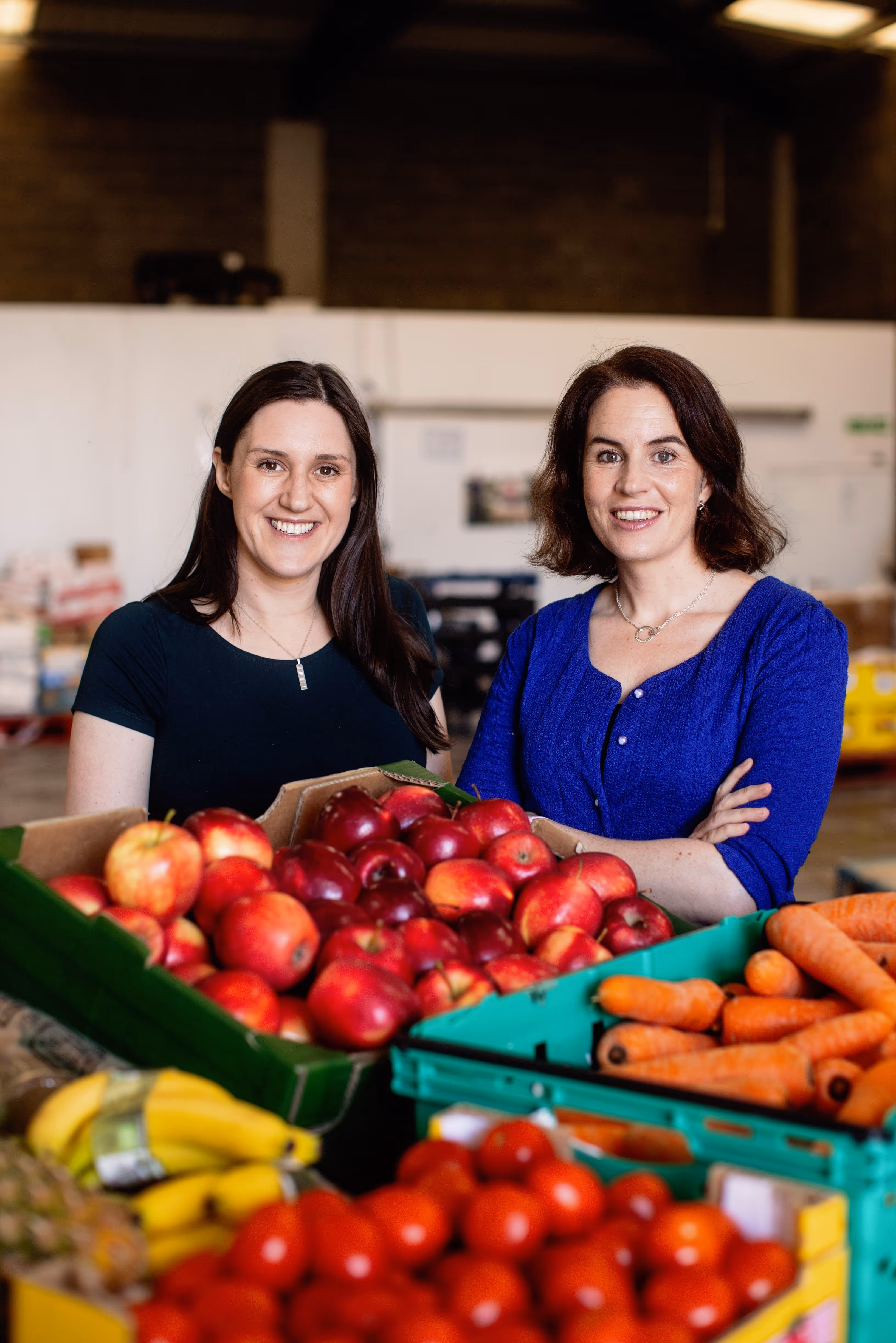 Two smiling women standing behind crates of fresh apples, carrots, bananas, and tomatoes in a warehouse setting.