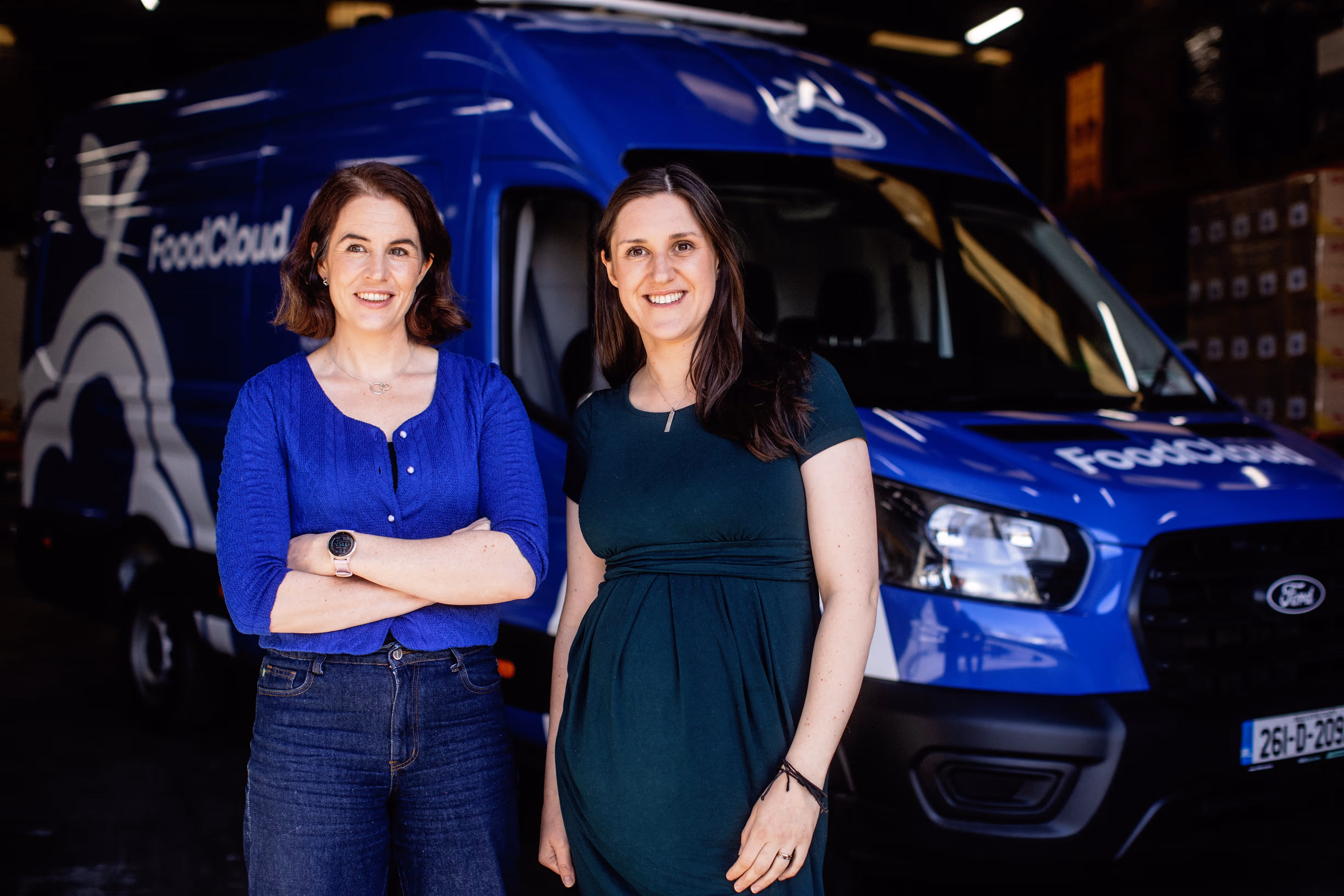 Two smiling women standing in front of a blue FoodCloud delivery van inside a warehouse.