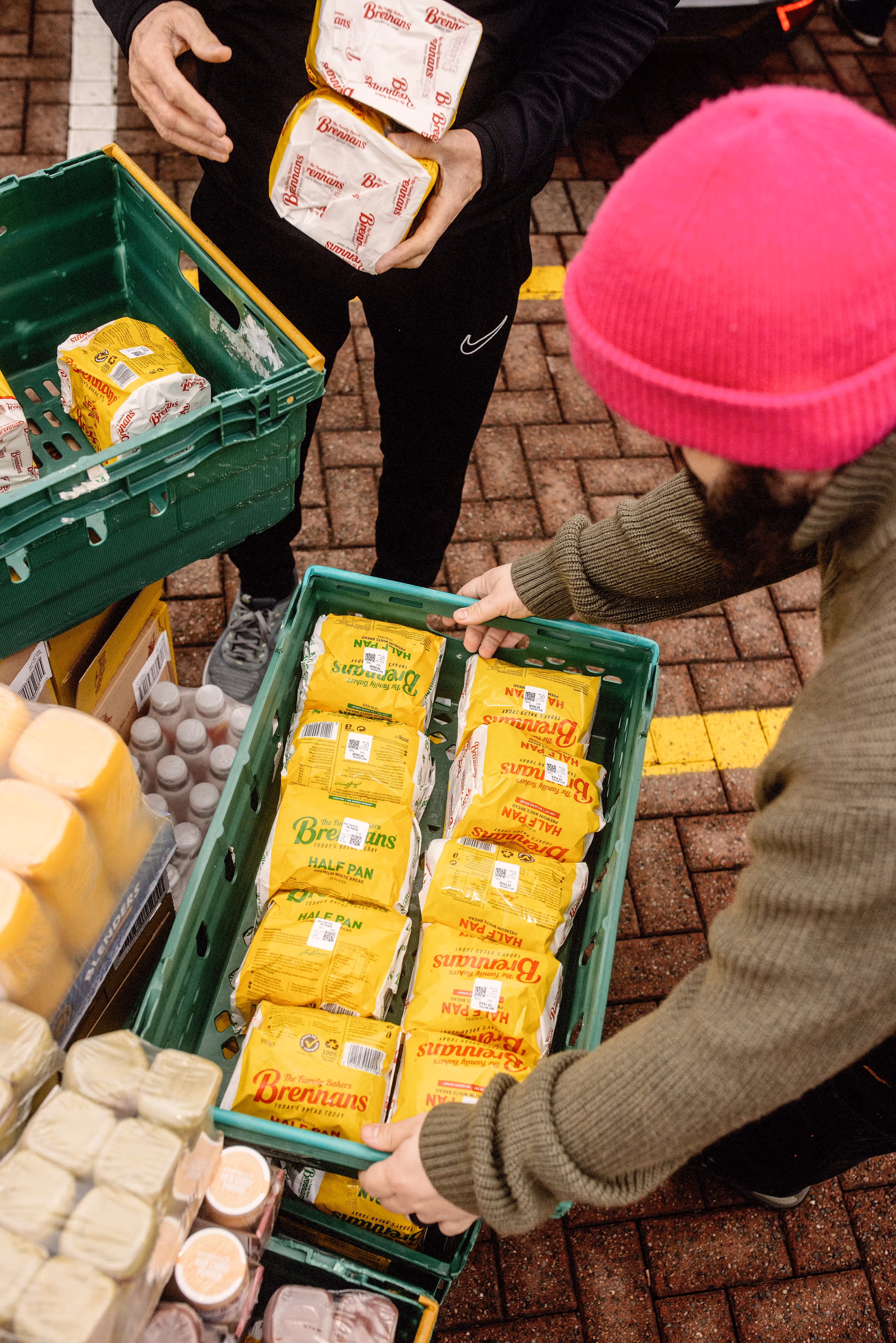 Two people handling green crates filled with yellow-packaged loaves of Brennan's bread on a brick pavement.
