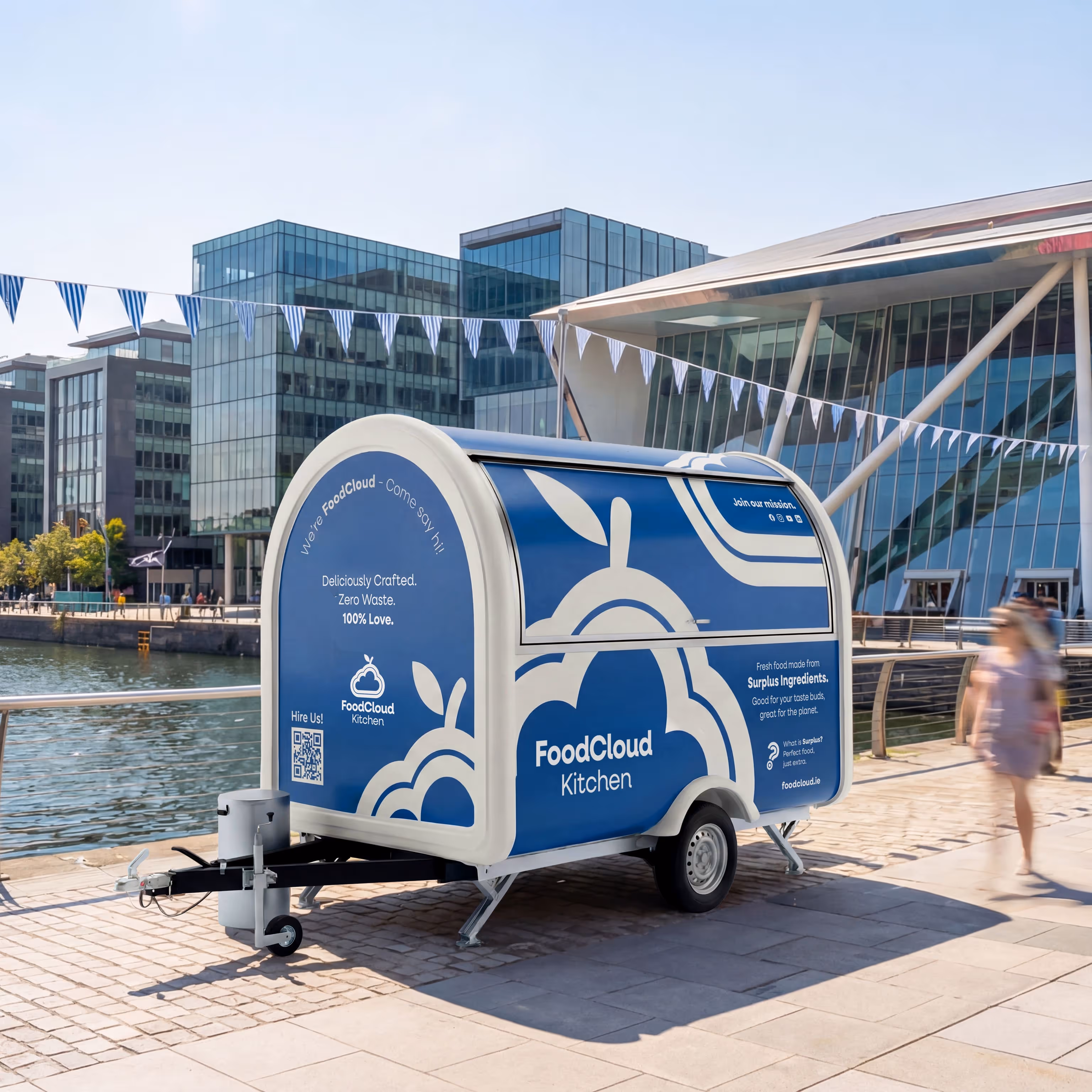 Blue and white FoodCloud Kitchen food trailer parked by waterfront with modern buildings and a pedestrian nearby.